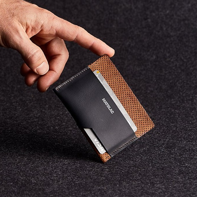 Close up of a man's finger balancing a black and brown genuine leather cash slip slim wallet on a grey background