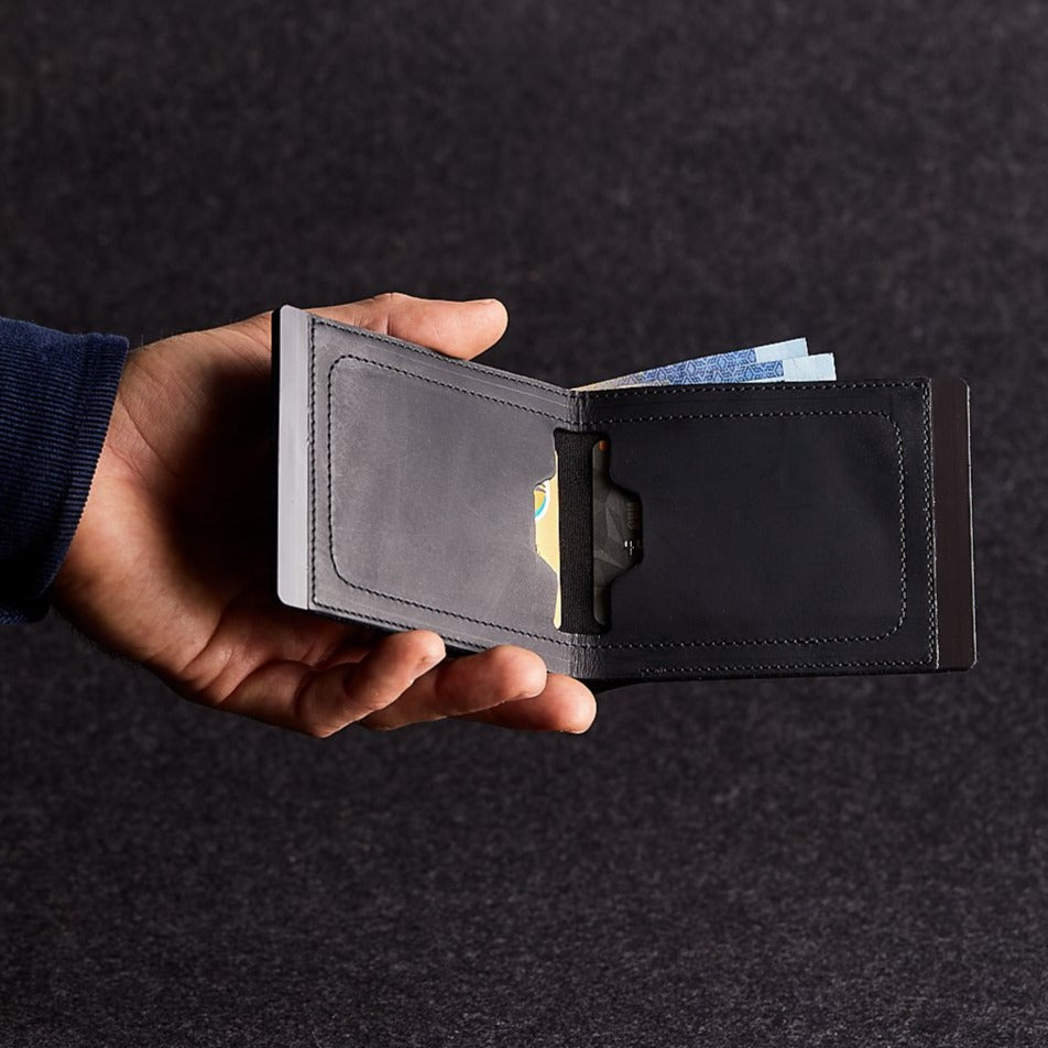 A man's hand holding open a minimalist black genuine leather wallet on a grey felt background