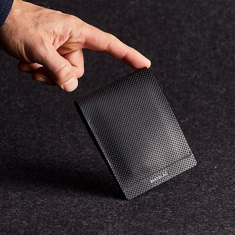 A man's hand balancing a minimalist black genuine leather wallet on a grey felt background